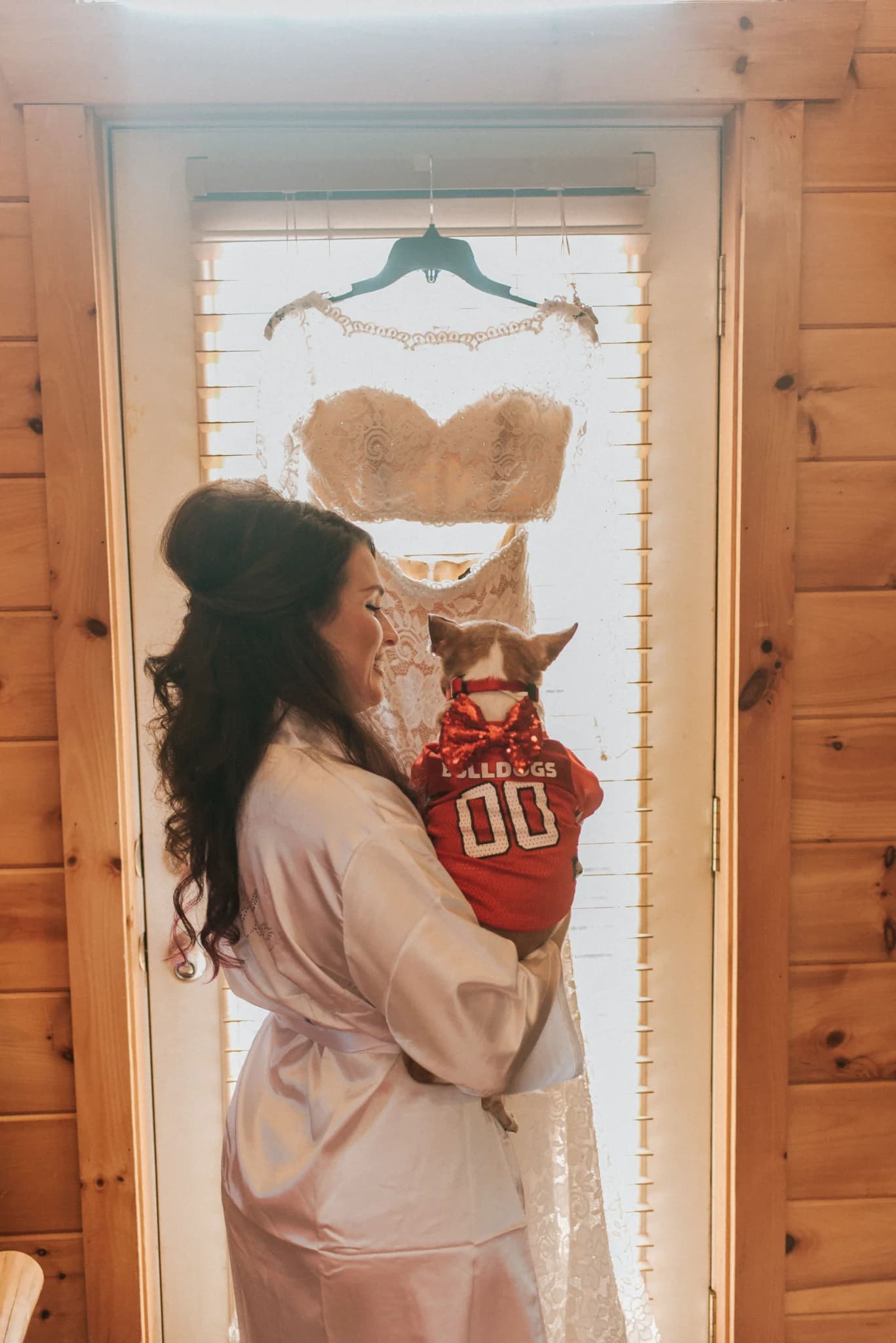 Bride in white robe holding dog with lace wedding dress hanging on door at Moose Hollow Lodge in Pigeon Forge