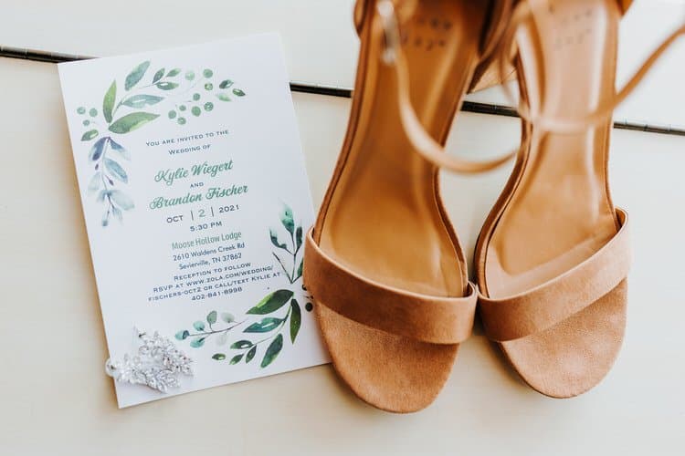 Wedding invitation for Moose Hollow Lodge, light brown sandals, and a silver hair clip on a light surface.