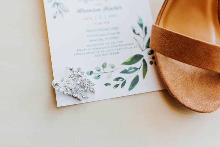Wedding invitation for Moose Hollow Lodge with leaf design, sparkling earrings, and a brown sandal on a light surface.