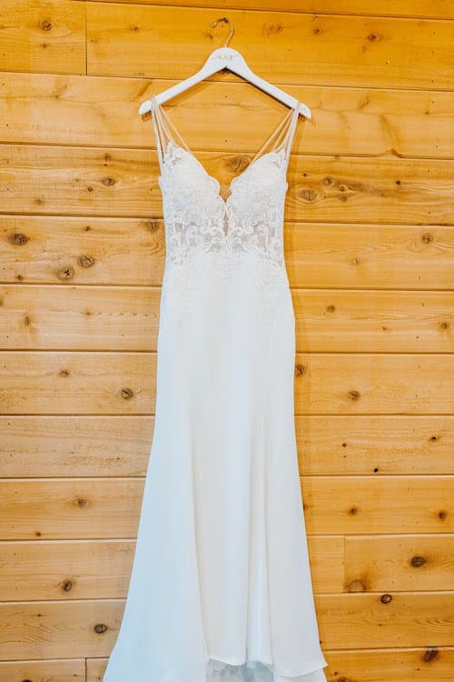White wedding dress with lace bodice and thin straps hangs on a personalized hanger against a wooden wall at Moose Hollow...