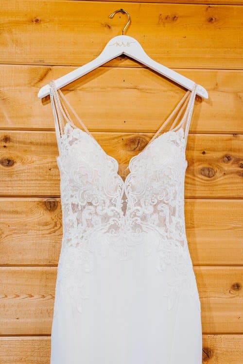 White wedding dress with lace top and delicate straps hanging on a custom 'mrs' hanger against a light wood wall.