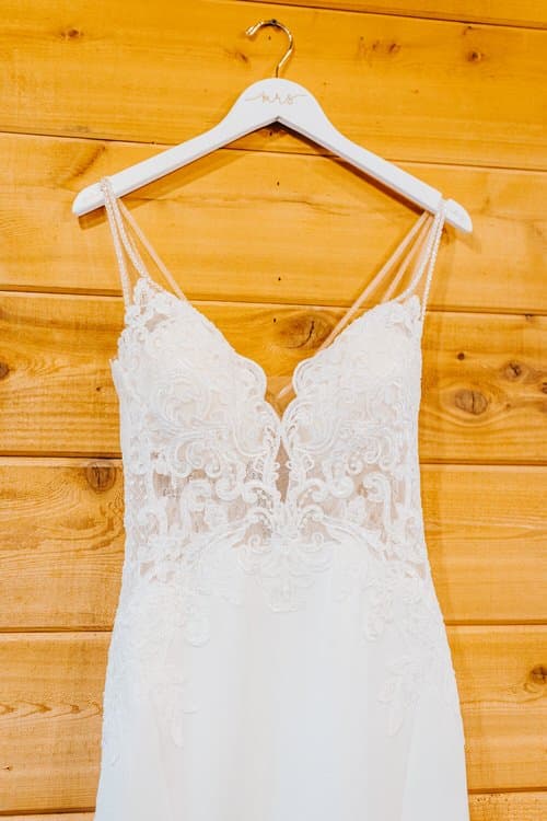 White lace wedding dress with delicate straps hanging on a 'mrs' hanger against a light wood plank wall.