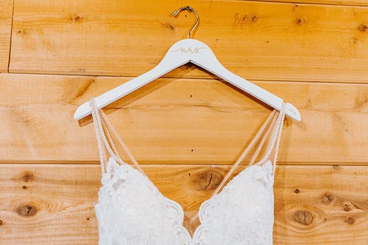 White lace wedding dress with beaded straps on a personalized 'Mrs.' hanger against a light brown wood plank wall.