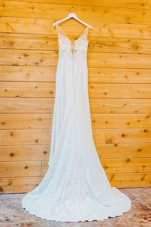 White wedding gown with lace and button back hanging on a white hanger against a rustic wood plank wall.
