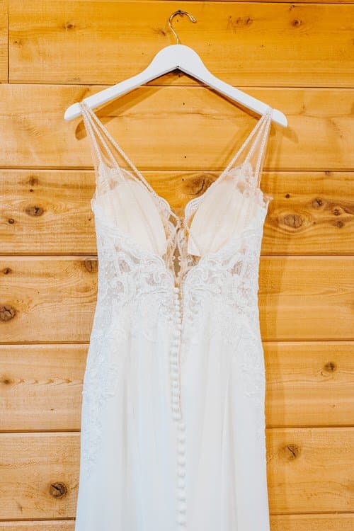 A white lace wedding dress with a button-up back hangs on a white hanger against a rustic wooden wall at Moose Hollow Lodge.