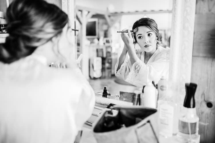 Woman in a lace-trimmed robe applies makeup in a mirror. Her reflection is visible, with wooden beams in the background...