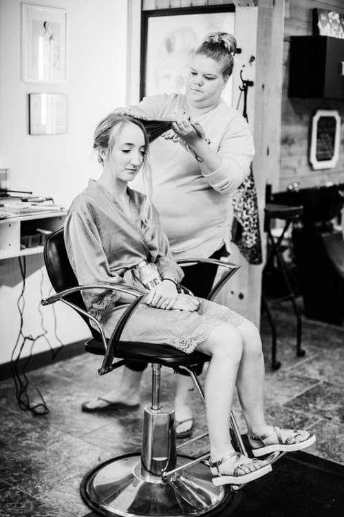 Woman in robe getting hair styled by a second woman in a salon chair. Black and white photo showing hair preparation.
