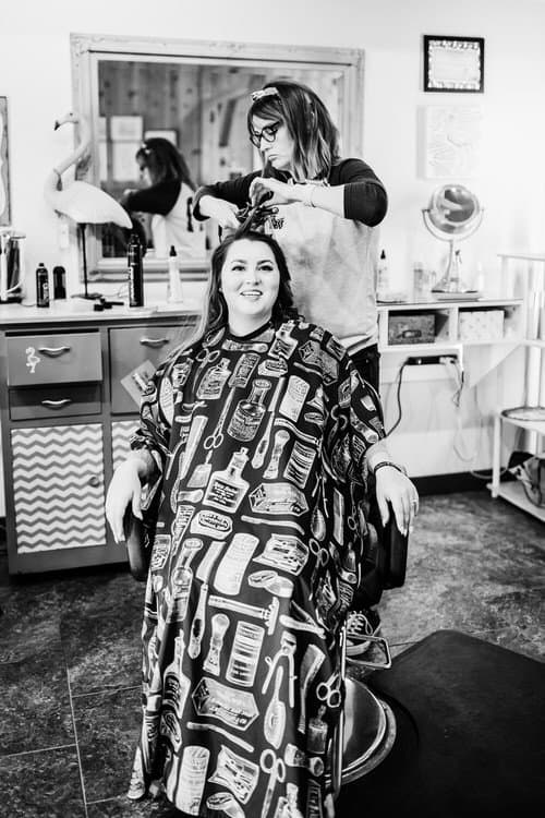 Woman in haircutting cape smiling while a stylist curls her hair in a bright room with a mirror and flamingo decor.
