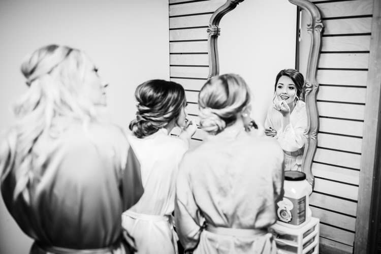 Three women in robes getting ready. One applies lipstick, reflected in an ornate mirror in a room with paneled walls.