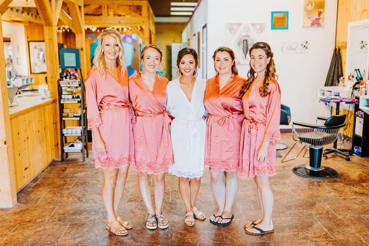 Five women, one in a white robe and four in pink robes, stand in a line indoors. A salon chair is visible.