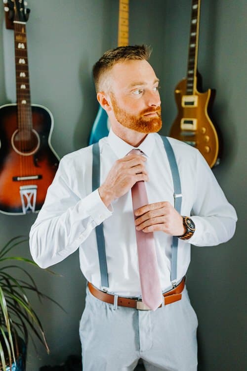 Man in white shirt, light grey suspenders, and pink tie adjusting his tie. Guitars hang on a grey wall in the background.