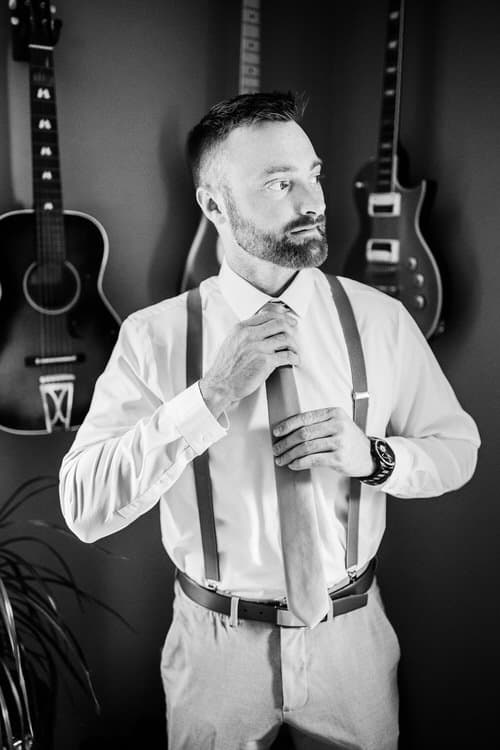 Man in white shirt, suspenders, and tie adjusts his tie. Acoustic and electric guitars hang on a dark wall behind him.
