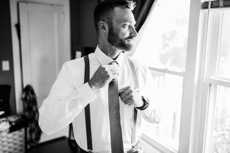 Black and white photo of a man in a white shirt and suspenders adjusting his tie while looking out a bright window.