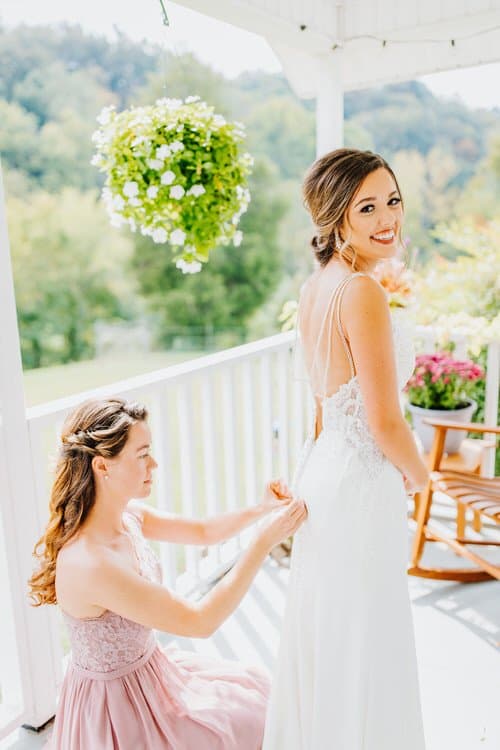 Person in pink dress buttons a white wedding gown on another person on a white porch with a hanging plant and green hills.