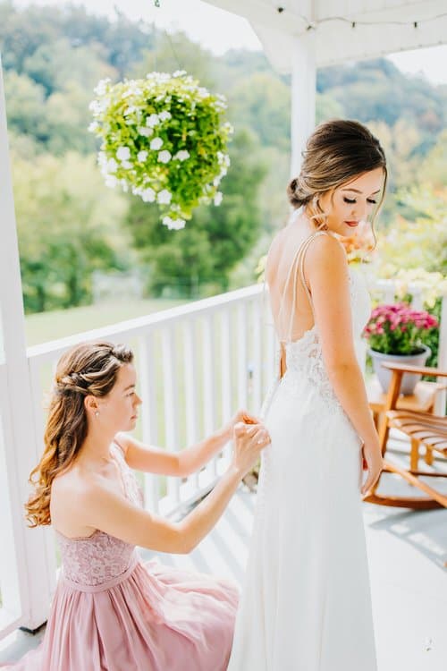 Woman in white wedding dress getting buttons fastened on a bright porch with green trees and hills in the background.