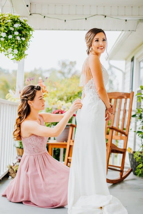Bride in white dress gets help with buttons from a bridesmaid in a pink dress on a porch with plants and a rocking chair.