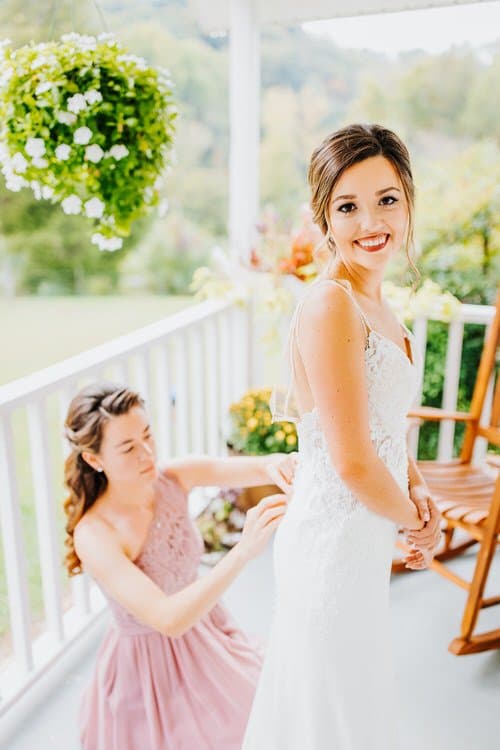 Bride in white lace dress smiling, another woman buttons her back on a white porch with flowers, Moose Hollow Lodge.