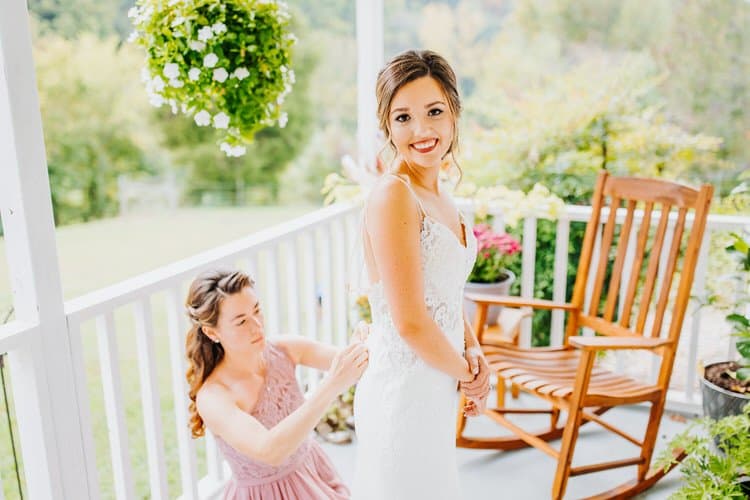 Bride smiles as bridesmaid fastens her white lace dress on a porch at Moose Hollow Lodge. Green trees visible.