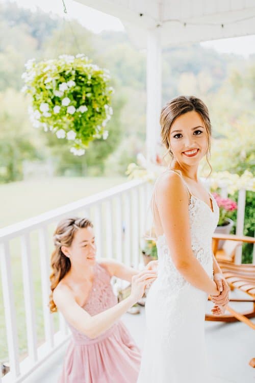 Bride in white lace gown smiles as attendant fastens dress on a covered porch with white railing and hanging flowers.