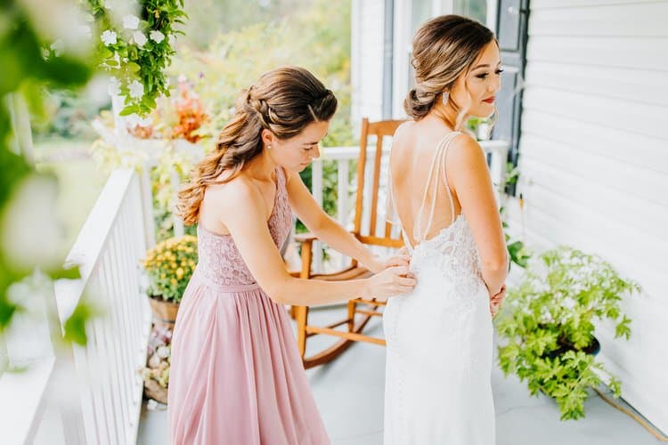 Woman in pink dress buttons back of wedding dress on another woman on a white porch at Moose Hollow Lodge, Sevierville.