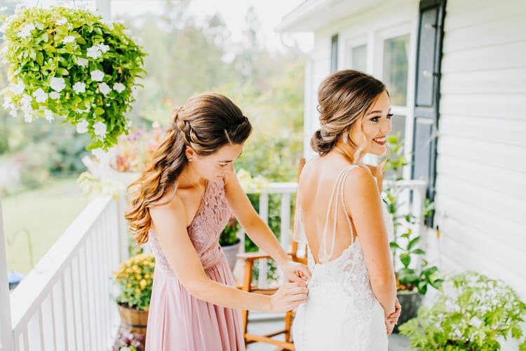 Bridesmaid helping a bride with her white lace wedding dress on a white porch railing. Hanging plants and white building...