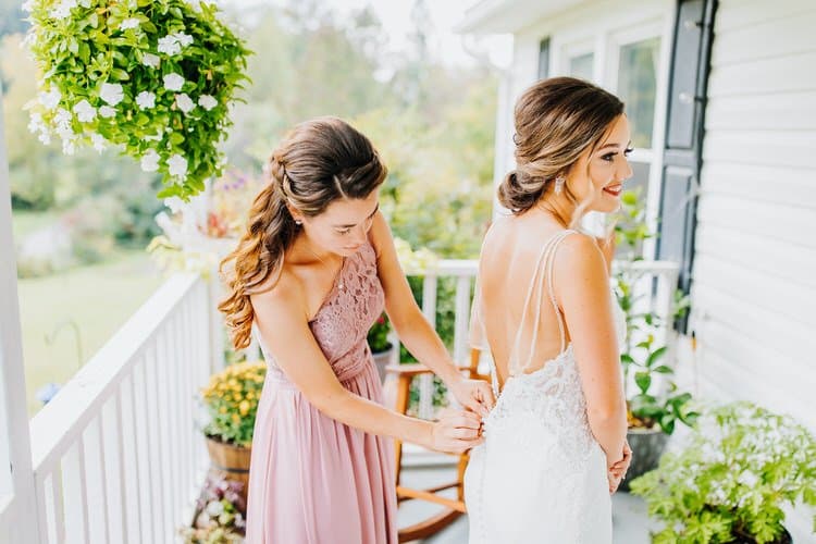 Person in pink dress zipping a bride's wedding gown on a porch with white railings and hanging plants at Moose Hollow Lodge.