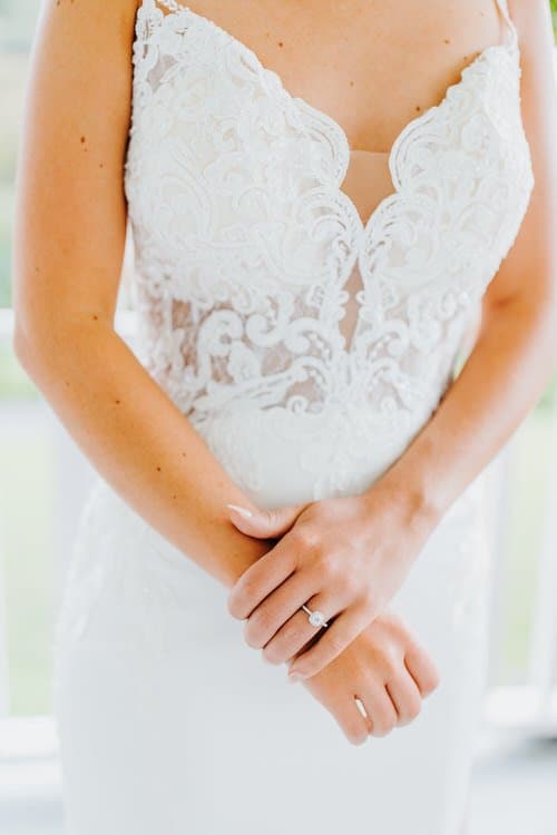 Close-up of a person in a white lace wedding dress, arms crossed. An engagement ring is visible on a finger.