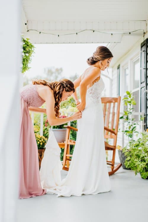 Bridesmaid in pink dress fastening buttons on a bride's white gown on a porch at Moose Hollow Lodge.