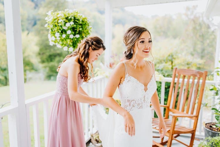 Bride in white dress on a porch, attendant adjusting her dress. Greenery and trees in background at Moose Hollow Lodge.