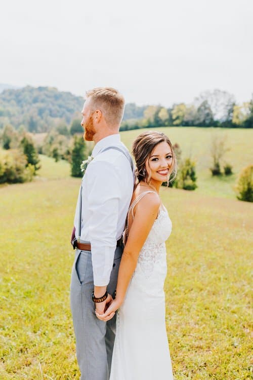 Bride and groom stand back-to-back, holding hands in a grassy field with rolling hills and trees under a clear sky.