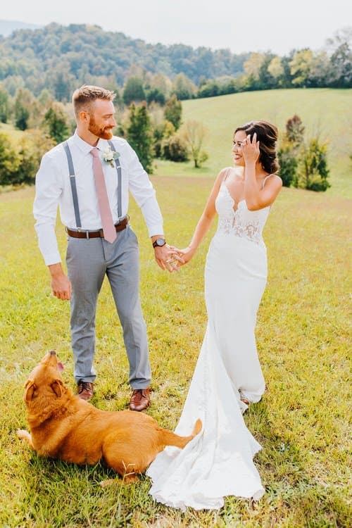 Wedding couple holding hands in a grassy field with a brown dog. Groom in suspenders, bride in white lace dress.