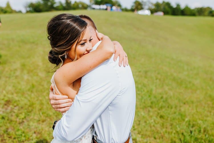 Bride and groom embracing in a sunny, open grassy field. Bride wears a white dress, groom a white shirt and brown belt.