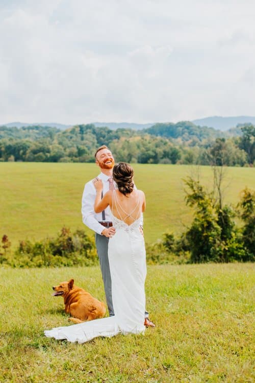 Bride in white dress, groom in suspenders, and a dog in a green field with mountains in the background at Moose Hollow Lodge.