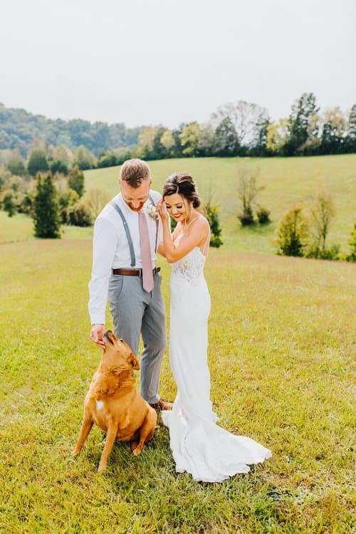 Wedding couple and golden dog in a field. Groom pets dog while bride smiles. Grassy hills and trees in background.