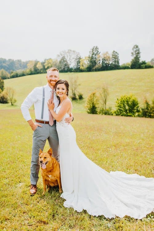 Wedding couple and their dog posing in a grassy field with rolling hills and distant trees under a bright sky.