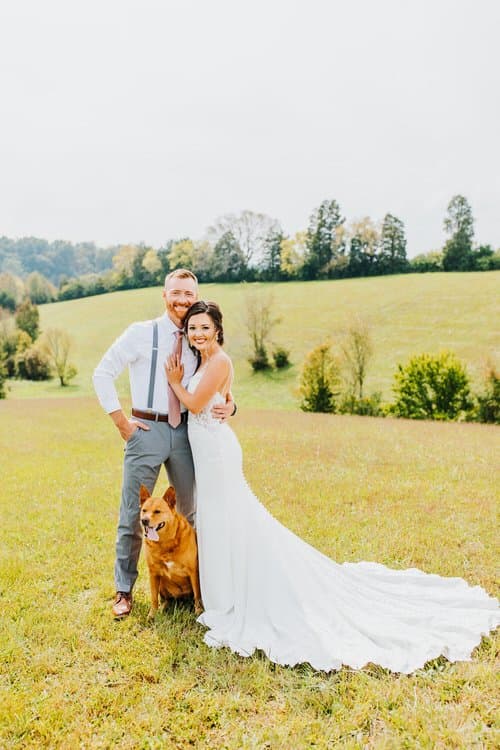 Wedding couple and dog stand in a grassy field with rolling hills and trees in the background. The bride wears a white gown.
