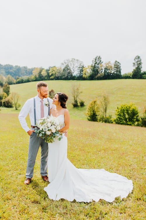 A bride in a white gown holding a large bouquet stands with a groom in a white shirt and suspenders in a grassy field.
