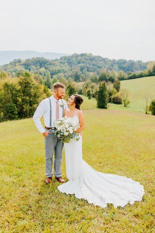 Bride and groom stand on a green grassy hill. Distant tree-covered mountains are visible under a clear sky. Outdoor...