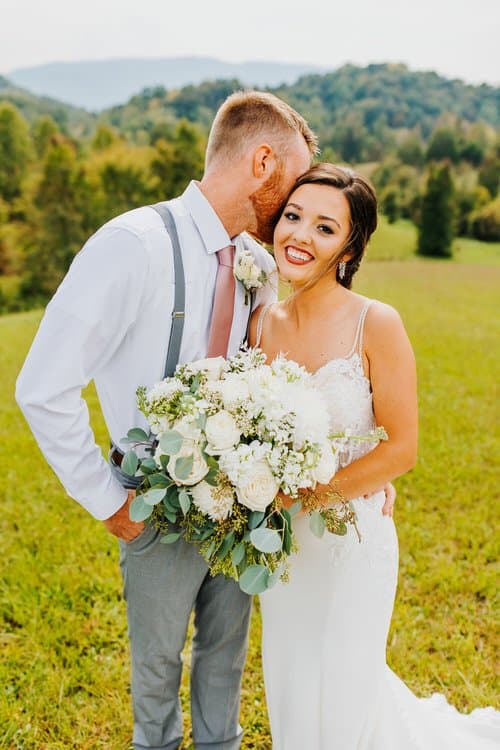 Bride and groom pose outdoors with a white floral bouquet, showing distant green mountains at Moose Hollow Lodge.