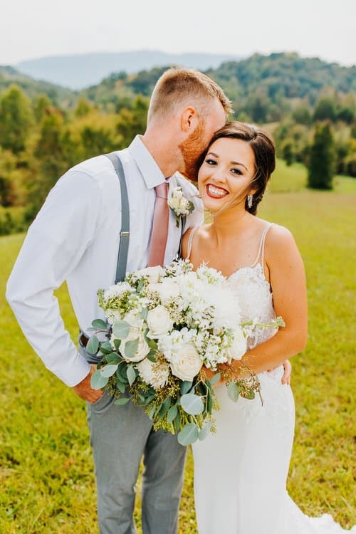 Bride in white dress holding bouquet, groom in suspenders kissing her head, standing in a green field with hills.