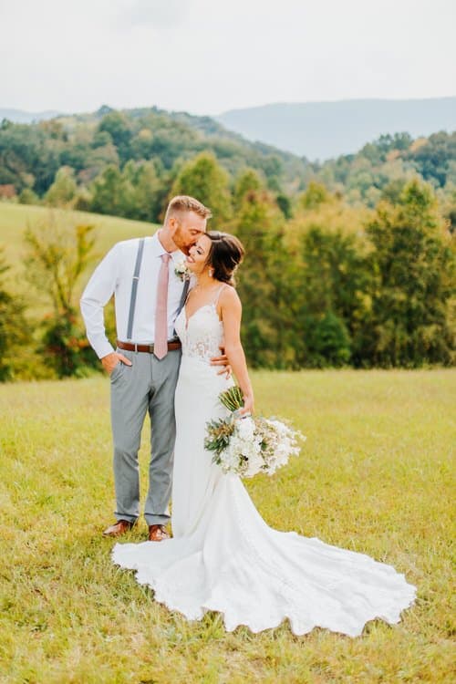 A wedding couple embraces at Moose Hollow Lodge with rolling Smoky Mountains in the background. Groom kisses bride's...