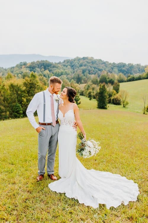Couple in wedding attire posing in a grassy field with forested hills in the background under a bright sky.