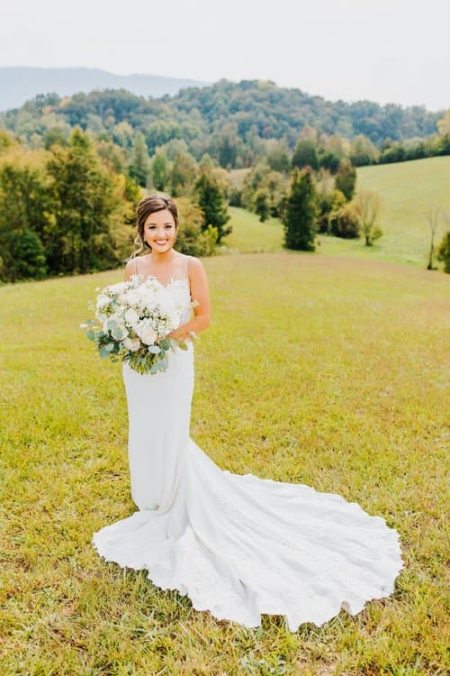 Bride in white gown holding a bouquet, standing in a grassy field with rolling tree-covered hills in the background under...