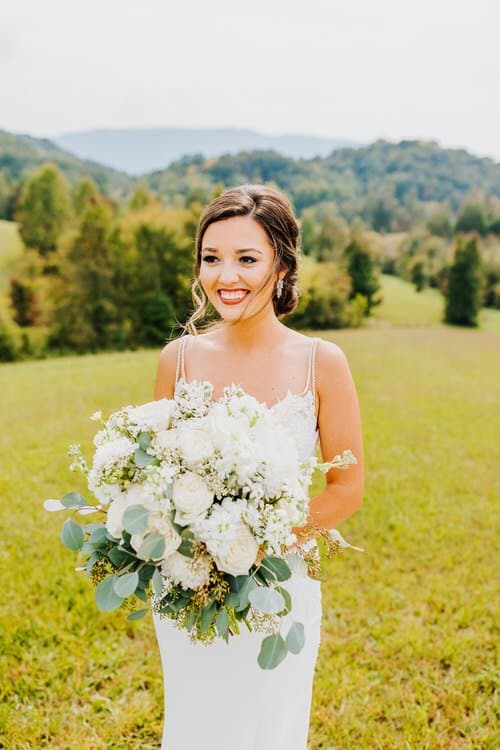 Bride in white dress holding large white and green bouquet in a grassy field with rolling hills and trees in the background.