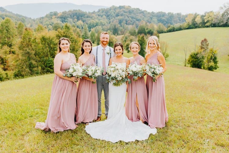 Bride, groom, and four bridesmaids posing outdoors at Moose Hollow Lodge with bouquets, against a backdrop of rolling...