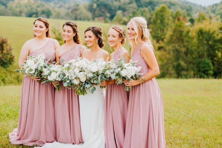 Five women, bride in white, four bridesmaids in mauve dresses, holding white and green bouquets, in a grassy field with...