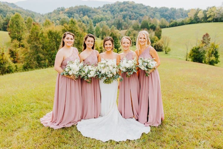 Bride in white gown with four bridesmaids in dusty rose dresses holding bouquets on a grassy hill with tree-covered...
