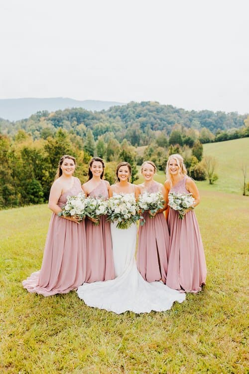 Bride in white gown with four bridesmaids in pink dresses holding bouquets in a grassy field, Smoky Mountains background.