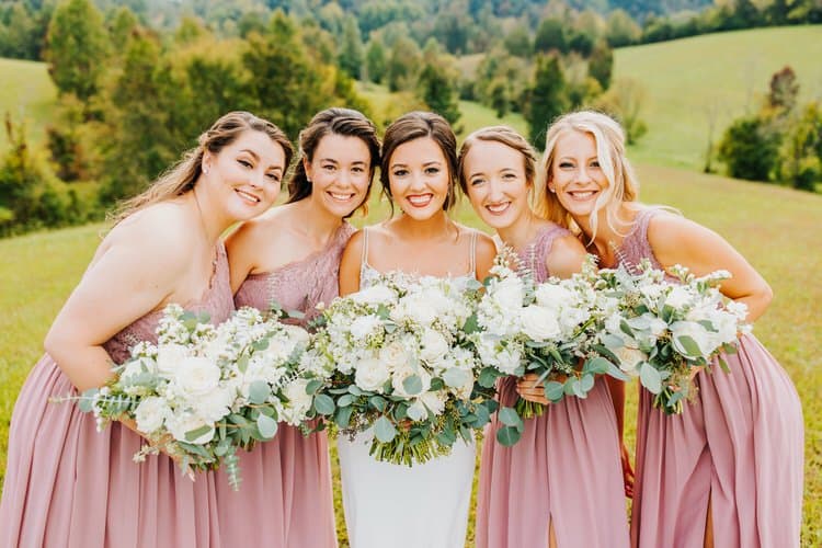 Five women in wedding attire, one bride, four bridesmaids, hold white and green bouquets in a field at Moose Hollow Lodge.