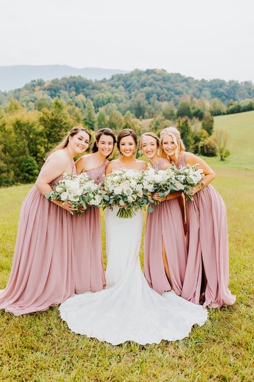 Bride and four bridesmaids in mauve dresses with white bouquets stand in a grassy field. Mountains are in the background.